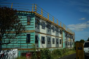 A new large three-story unfinished housing block with eighteen windows on each of the second and third floors overlooks an old unpainted natural wooden fence covered in overgrown weeds and grass and a neighbouring concrete driveway. The facing exterior walls have light green building paper and steel scaffolding which is also on the roof.